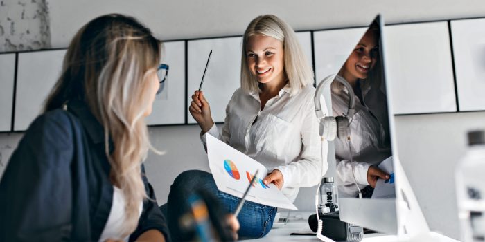 Smiling blonde female manager holding infographic and pencil, while sitting on table. Indoor portrait of two girls working with computer in office.. Smiling blonde female manager holding infographic and pencil, while sitting on table. Indoor portrait of two girls working with computer in office.
